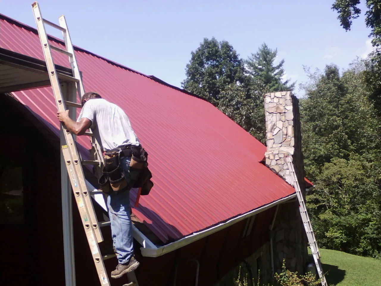 Roofer on a ladder beside a red metal roof and stone chimney — Soffit & Fascia Repair by Roofing Brothers in Tenino
